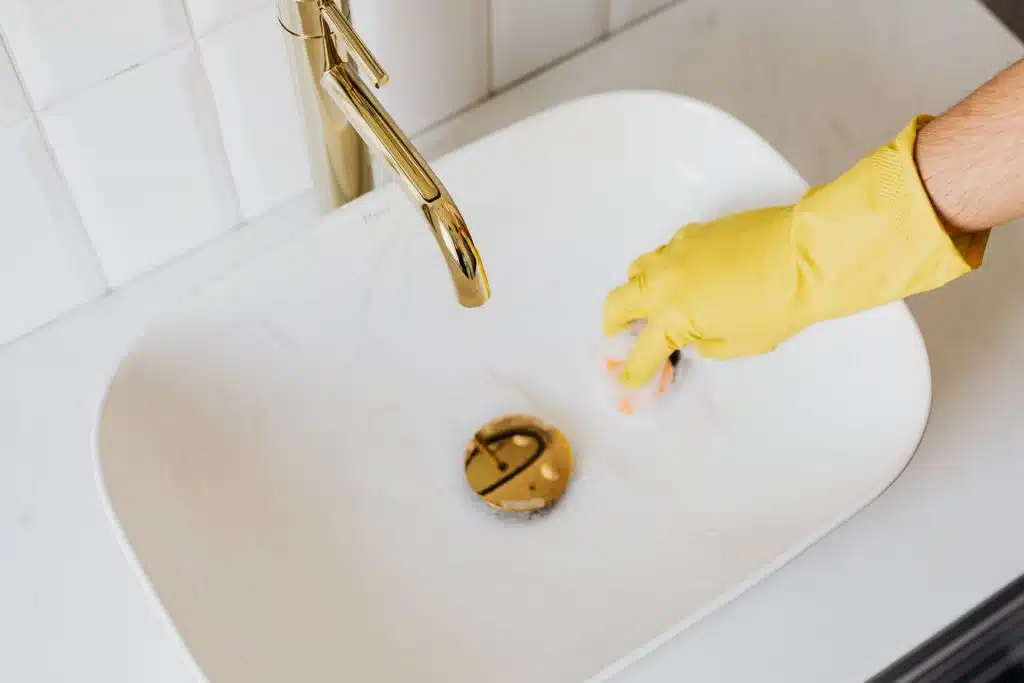 Hand in yellow rubber glove cleaning a modern white porcelain bathroom sink near a brass gold faucet.