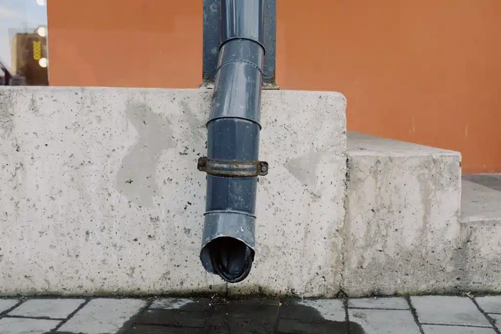 Dark gray metal gutter downspout mounted against a concrete wall base over paved sidewalk.